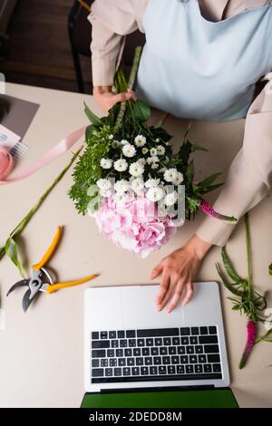 Cropped view of female florist using laptop, while holding bouquet on desk with secateurs and decorative ribbon Stock Photo