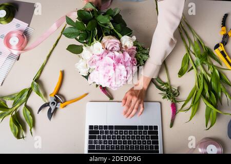 Cropped view of female florist using laptop, while holding bouquet on desk with tools, plants and decorative ribbons Stock Photo