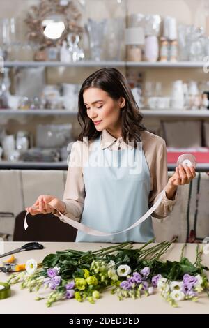 florist in apron holding decorative ribbon near eustoma flowers on table near rack with vases on blurred foreground Stock Photo