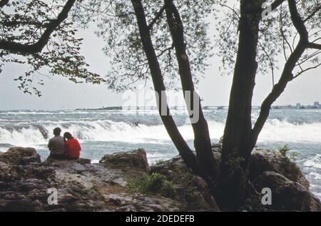 1970s Photo (1973) -  Couple enjoy view of Niagara River rapids from the shore of Goat Island which separates the American and Canadian falls at the brink of the cataract Stock Photo