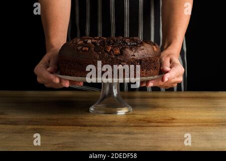 Woman holding a cake stand with a chocolate cake on a wooden table Stock Photo