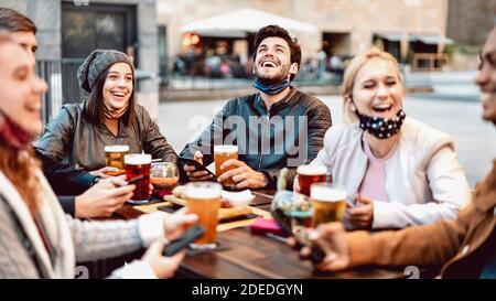 Young friends drinking beer wearing face mask - New normal lifestyle concept with people having fun together talking on happy hour at outside brewery