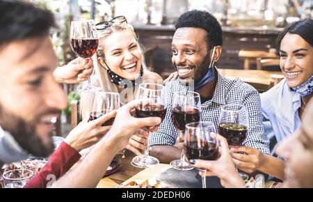 Friends toasting red wine at outdoor restaurant bar with open face mask - New normal lifestyle concept with happy people having fun together