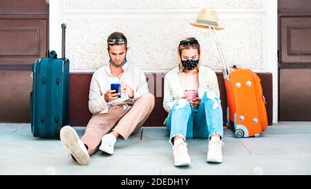 Young couple with face mask using mobile smart phones - Young people checking online reservations sitting on floor outside train station Stock Photo