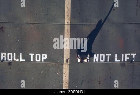 1970s Photo (1973) -   Camera points directly down on the elongated shadow of a man standing on the roof of a great barge in the Houston Ship Channel. The man is signalling directions to a towboat which is guiding the barge along the channel Stock Photo