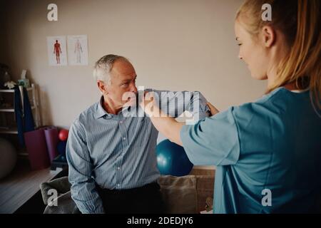 Young female physiotherapist examining shoulder and elbow of senior patient in hospital room Stock Photo