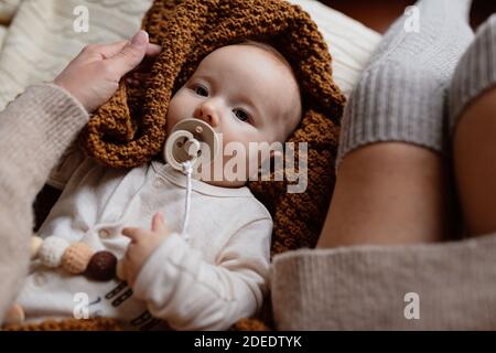 Cute caucasian baby girl lying on cozy bed with her mother dressed in knitted sweater brown color. Stay at home during Coronavirus covid-19 quarantine Stock Photo