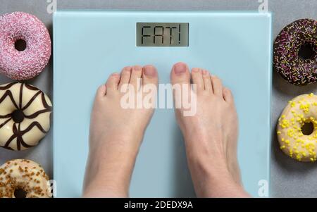 Person standing on digital weight scale showing the words FAT, and junk food donuts on background, diet concept Stock Photo