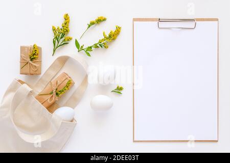 Happy easter concept. White eggs, yellow flowers, craft gifts fly out textile bag and clipboard with paper on white background Mockup Flat lay Top vie Stock Photo