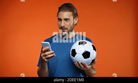 surprised football fan in blue t-shirt using smartphone and holding soccer ball on orange Stock Photo