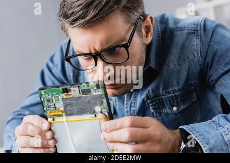 Focused repairman in eyeglasses looking at part of broken digital tablet Stock Photo