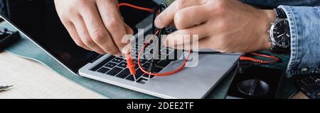 Cropped view of repairman holding sensors of multimeter on broken keyboard of laptop at workplace, banner Stock Photo