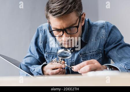Focused repairman in eyeglasses with magnifier looking through at broken laptop with blurred workplace on foreground Stock Photo