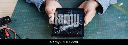 Cropped view of repairman holding smashed digital tablet at workplace, banner Stock Photo