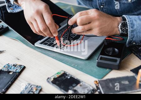 Cropped view of repairman holding sensors of multimeter on broken keyboard of laptop with blurred disassembled phones on foreground Stock Photo