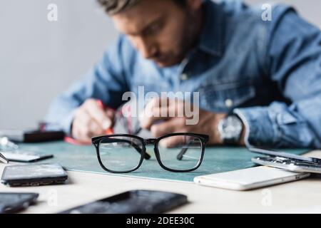 Eyeglasses on table near disassembled parts of mobile phones with blurred repairman working on background Stock Photo