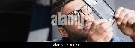 Focused repairman in eyeglasses looking at disassembled part of broken mobile phone on blurred background, banner Stock Photo