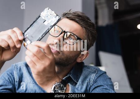Focused repairman in eyeglasses looking at disassembled component of mobile phone on blurred background Stock Photo