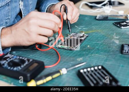 Cropped view of repairman holding multimeter sensors on disassembled part of mobile phone on blurred foreground Stock Photo