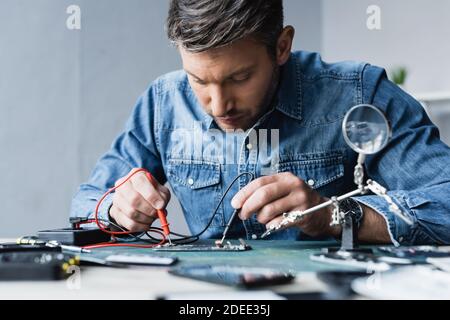 Focused repairman holding sensors of multimeter on disassembled part of mobile phone on blurred foreground Stock Photo
