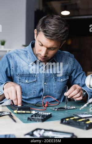 Focused repairman regulating multimeter while holding sensor on disassembled part of mobile phone on blurred foreground Stock Photo
