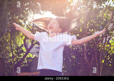 Happy young girl alone, little cheerful school age child jumping up, flying dispelled hair, arms spread, sunlight, outdoors portrait. Childhood Stock Photo