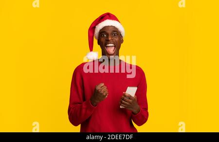 Excited Black Guy In Santa Claus Hat Celebrating Success With Smartphone Stock Photo