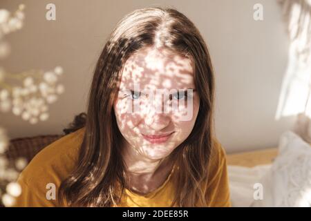 Portrait of young beautiful woman by the window with shadow from flowers on her face. Morning spring aesthetics Stock Photo