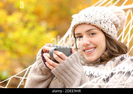 Happy woman holding coffee cup lying on hammock looking at camera in autumn holiday Stock Photo