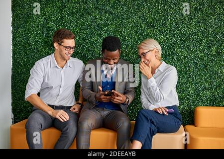 three diverse colleagues sit talking having fun apart form work, laugh looking videos on smartphone Stock Photo