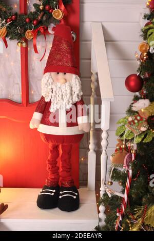 A figurine of red Santa Claus stands near a red door and a Christmas tree inside the house Stock Photo
