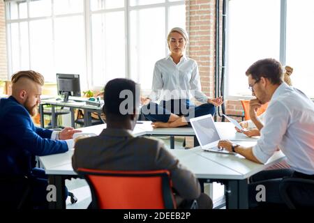 young caucasian woman meditating in office while colleagues are working, keep calm, meditate. sits on table with crossed legs Stock Photo