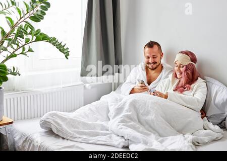 man and woman have rest with smartphone on bed, they lie on bed in the morning at weekend, caucasian man and woman watching video or something else to Stock Photo