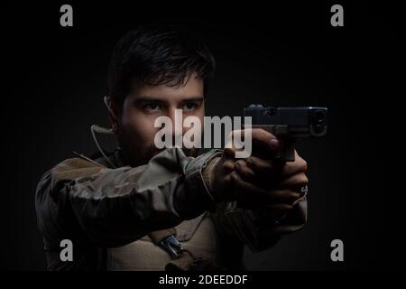 closeup to a Latin man with a beard standing, holding a gun, wearing a headphones and camouflage clothing, studio Stock Photo