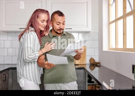 couple with papers in the kitchen, young caucasian married couple checking bills documents in the kitchen, discuss Stock Photo