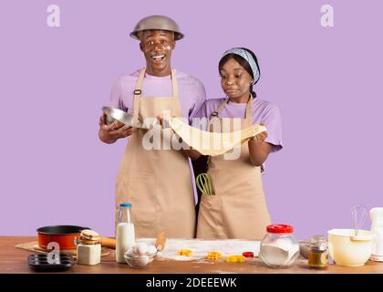 Positive black couple in aprons having dirty faces, holding cookie dough, baking together on lilac studio background Stock Photo