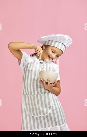 smiling little girl in chef hat and apron mixing something with a whisk in bowl over pink background. child dreams of becoming a chef Stock Photo