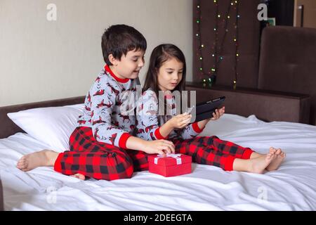 A boy and a girl in pajamas are sitting at home on the bed, looking in surprise at the digital tablet. Stock Photo