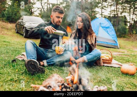 Man pours his girlfriend tea from thermos while sitting in the nature near campfire Stock Photo