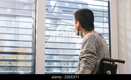 Man on wheelchair in living room looking through window. Handicapped invalid paralysed person, depressed alone at home. Stock Photo