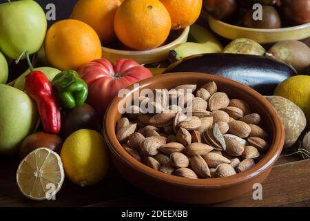 A homemade clay bowl filled with whole almonds in their shell on an old wooden table with fruits, vegetables, and organic and natural ingredients. Stock Photo