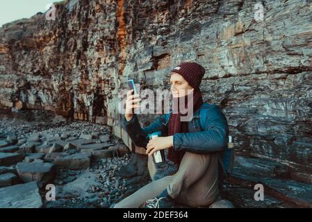 A young man in warm clothes sitting on the stone and taking pictures of the sunset on his cell phone camera. Rest during a walk on the rocky coast. Lo Stock Photo