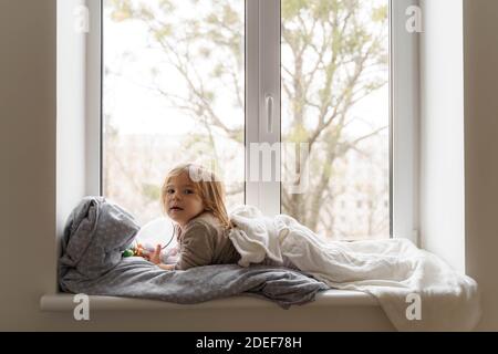 Child lying on the window in living room and watching at the street. cozy home on weekends, holidays and lockdown Stock Photo