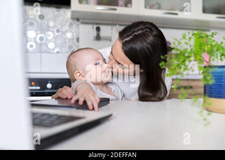 Portrait of mom and baby son sitting at home in kitchen, loving mother kissing baby Stock Photo