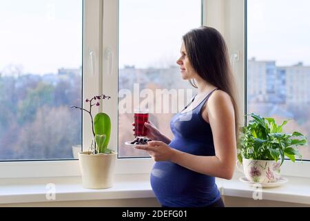 Young beautiful pregnant woman drinking berry juice from glass at home near window Stock Photo