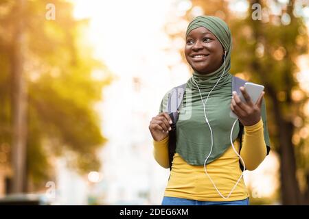 Smiling black muslim woman in headscarf walking by street and listening music on smartphone Stock Photo