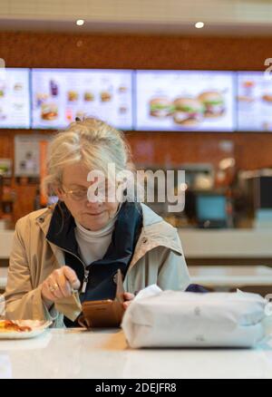 In the cafeteria an old woman sitting and using her smartphone while having her breakfast. Selective focus, street view. Stock Photo