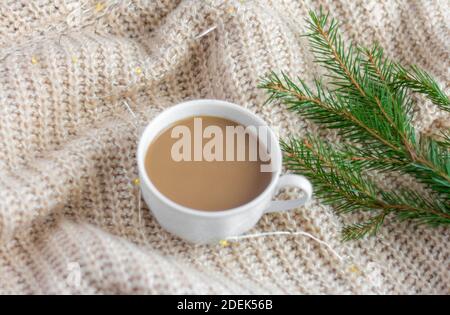 A Cup of hot coffee with milk on a warm blanket with a garland and a sprig of spruce.Christmas concept.spruce branch on a woolen blanket. Stock Photo