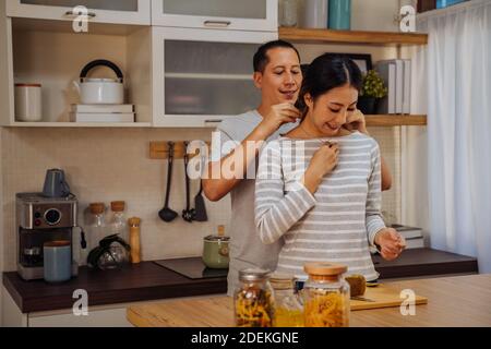 Young man giving his girlfriend a necklace as present. Asian woman getting surprised by her boyfriend with marriage proposal while cooking in kitchen at home Stock Photo