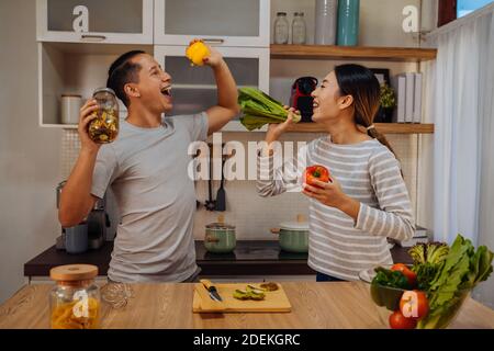 Young lovely couple dancing in the kitchen with window. Asian woman and Caucasian man singing with vegetables and ingredients in modern home Stock Photo